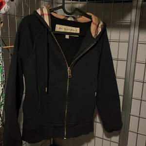 Säljer en snygg svart hoodie från Burberry med klassiskt rutigt mönster i luvan. Den har dragkedja framtill och är i mycket bra skick. Perfekt för höst och vår, eller när du vill ha en stilren look. Passar både till vardags och lite finare tillfällen. Är en dam kofta.