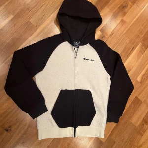 Svart och vit hoodie från Champion - Mysig fleecehoodie från Champion i svart och vitt. Den har en dragkedja framtill och en praktisk huva. Perfekt för kyliga dagar med sin varma och sköna känsla. Jackan har långa ärmar och en broderad logga på bröstet. Perfekt för höst och vinter!