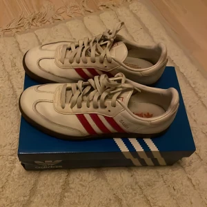 Adidas Samba i vitt och rött - Säljer ett par klassiska Adidas Samba sneakers i vitt med röda ränder. Skorna är i bra skick. Perfekta för både vardag och träning. Storlek och passform är normal.