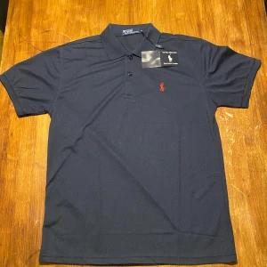 Mörkblå pikétröja från Ralph Lauren - Säljer en snygg mörkblå pikétröja från Ralph Lauren i nytt skick. Den har en klassisk krage med knappar och det ikoniska röda broderade logotypen på bröstet. Perfekt för både vardag och festliga tillfällen. Tröjan är kortärmad