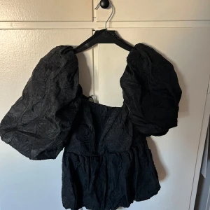 Svart blus med puffärmar - Säljer en elegant svart blus med stora puffärmar och vacker broderad textur. Perfekt för festliga tillfällen eller en utekväll. Blusen har en smickrande passform och en djup urringning. Öppen i ryggen med knytning. Den är i nytt skick med etikett kvar.