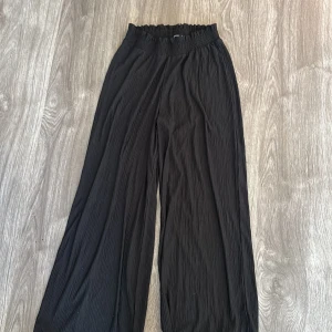 Svarta vida byxor i kortare modell - Säljer ett par svarta vida byxor i mycket bra skick. De är högmidjade med en elastisk midja för extra komfort. Materialet är lätt och luftigt, perfekt för både vardag och fest. Byxorna har en ribbad struktur som ger en snygg detalj. Passar perfekt till både sneakers och klackar!