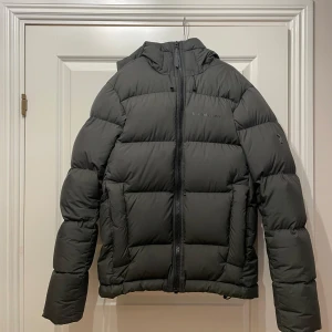 Grön pufferjacka från Peak Performance - Säljer en snygg grön pufferjacka från Peak Performance i mycket bra skick. Jackan har en dragkedja framtill och en mysig huva. Perfekt för kalla vinterdagar och håller dig varm och stilren. Passar både till vardags och utomhusaktiviteter. En innerficka finns och två ytterfickor. Går att ändra passformen på huva och jackans midia. Nypris 4200kr. 