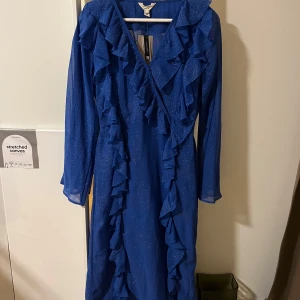 Blå glittrig klänning med volangdetaljer - Säljer en superfin blå klänning med glittrigt tyg och volangdetaljer. Den har en omlott-design och långa ärmar, perfekt för festliga tillfällen. Klänningen är i nytt skick och har en elegant v-ringning. 