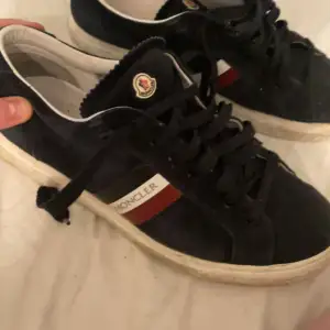 Säljer ett par snygga svarta Moncler sneakers i bra skick. De har en klassisk design med snörning och Monclers logga på sidan. Skorna har en vit sula och detaljer i rött och vitt på sidan. Perfekta för både vardag och fest! Pris är inte hugget i sten.