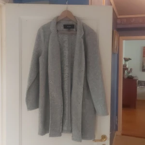 Grå lång kappa - Säljer en stilren och mysig grå lång kappa. Perfekt för höst och vinter med sin långa design och knappar framtill. Den är i ett mjukt material som känns skönt mot huden. Passar både till vardags och lite finare tillfällen.