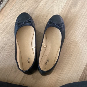 Svarta ballerinaskor med rosett - Säljer ett par svarta ballerinaskor från Alley i storlek 37. De är i syntetmaterial och har en söt rosett på tån. Perfekta för både vardag och fest!