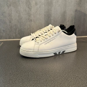 Vita sneakers från Emporio Armani - Säljer ett par stilrena vita sneakers från Emporio Armani. Skorna har en klassisk design med svart detalj vid hälen och märkets logga på sidan och sulan. Bra skick och Pris kan diskuteras!