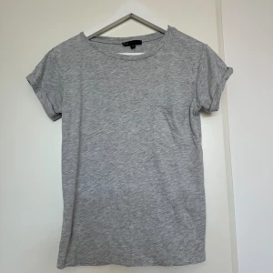Grå t-shirt från Moss Copenhagen - Säljer en stilren grå t-shirt från Moss Copenhagen i storlek XS. Den är tillverkad av 100% viskos vilket gör den supermjuk och skön att ha på sig. T-shirten har korta ärmar och en liten bröstficka som ger en extra touch. Perfekt för vardagsbruk!