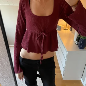 Vinröd ribbad topp - Säljer en vinröd ribbad topp med långa ärmar och knytning i midjan. Toppen har en croppad design och en rundad halsringning. Perfekt för en stilren look.