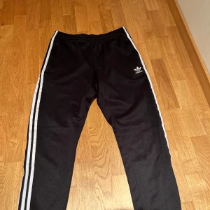 Adidas sst  - Snygga svarta träningsbyxor från Adidas med klassiska vita ränder längs sidorna. Byxorna har en elastisk midja och en liten Adidas-logga på ena benet. Perfekta för träning eller en avslappnad stil. Helt nya aldrig använt bara vid test köpt från footlocker vid flera bilder så kom privat så löser vi det !