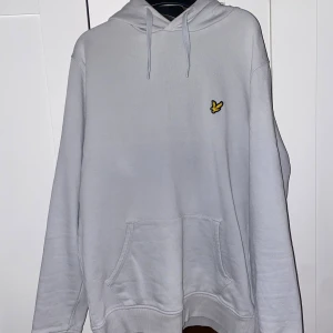 Grå hoodie från Lyle & Scott - Säljer en stilren grå hoodie från Lyle & Scott med deras ikoniska logga på bröstet. Tröjan har en klassisk känguruficka och justerbar huva med dragsko. Perfekt för en avslappnad look.