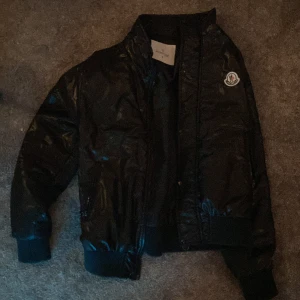 Svart pufferjacka från Moncler - Snygg svart pufferjacka från Moncler med glansig finish och ikonisk logga på bröstet. Jackan har en praktisk ficka med knapp på ärmen och dragkedja framtill. Perfekt för kyligare dagar.