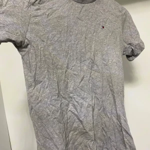 Grå t-shirt från Tommy Hilfiger - Säljer en klassisk grå t-shirt från Tommy Hilfiger med en liten logga på bröstet. T-shirten är kortärmad och tillverkad i mjukt bomullsmaterial. Perfekt under sommaren och för en avslappnad stil.