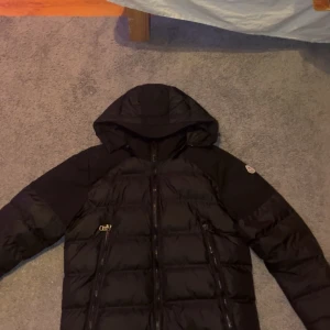 Moncler jacka  - Säljer en snygg svart dunjacka från Moncler. Jackan har en praktisk huva och dragkedja framtill. Perfekt för kyliga dagar och håller dig varm och stilren. Passar bra till både jeans och träningsbyxor. Jackan är storlek 2 som motsvarar S. Jag är 175cm lång och väger ca  70 kg och storlek S passar mig bra. Den passar perfekt. Hör gärna av er om ni har funderingar eller vill ha mer bilder på jackan. 
