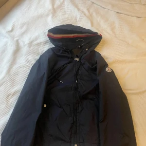 Mörkblå vindjacka från Moncler - Säljer en snygg mörkblå vindjacka från Moncler med huva och dragkedja. Jackan har en randig detalj i rött och vitt på huvan och Moncler-loggan på ärmen. Perfekt för blåsiga dagar!