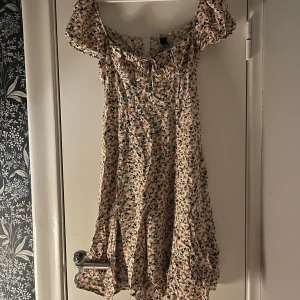 Blommig klänning med korta ärmar - Blommig klänning med korta ärmar, en slits och en söt knytning framtill. Perfekt för sommarens varma dagar. Den har en lätt och luftig design med ett vackert blommönster i mjuka färger.