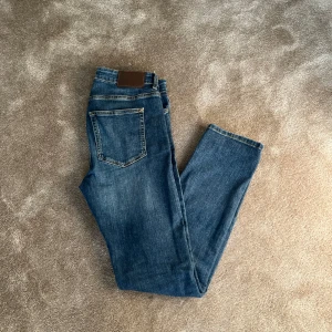 Blå jeans byxor  - Snygga blå jeansbyxor med klassisk femficksdesign och en bekväm passform. De har en knapp och dragkedja framtill samt en läderdetalj vid midjan. Perfekta för en avslappnad stil. Det är w28 och l30