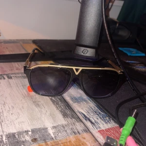 Solglasögon från Louis Vuitton - Snygga solglasögon från Louis Vuitton med en stilren design. Bågarna är i guld och svart med mörka linser. Perfekta för att ge en lyxig touch till din stil.