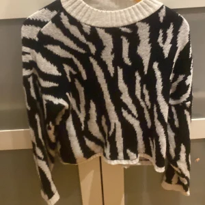 Svart och vit stickad tröja från Malady - Säljer en cool stickad tröja från Malady med ett zebramönster i svart och vitt. Tröjan har en croppad passform och långa ärmar. Perfekt för att ge din outfit en trendig touch!