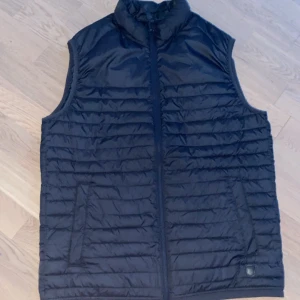Svart quiltad väst från Jack & Jones - Säljer en svart quiltad väst från Jack & Jones i storlek L. Västen har en dragkedja framtill och är perfekt för lager-på-lager under kyligare dagar men funkar utmärkt för sommar dagar också. Den är lätt och smidig att bära.