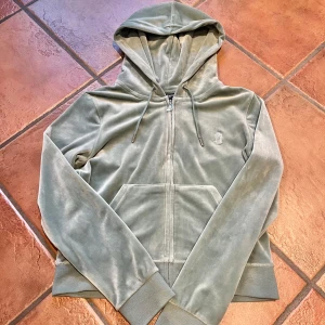 Grön hoodie från Juicy Couture - Säljer en mjuk och stilren grön hoodie från Juicy Couture. Den har en dragkedja framtill, långa ärmar och en praktisk huva med dragsko. Perfekt för en avslappnad look.