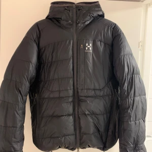 Haglöfs Dunjacka - Säljer en svart dunjacka från Haglöfs i storlek M. Jackan har en dragkedja framtill och en praktisk huva. Perfekt för kyliga dagar med sin quiltade design som håller värmen. Två sidofickor med dragkedja för säker förvaring.