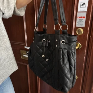 Svart quiltad axelväska - Snygg svart axelväska med quiltat mönster och dragkedja. Väskan har dubbla handtag och dekorativa detaljer i form av snörning och metallringar. Perfekt för att ha till skolan