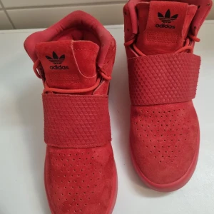 Röda sneakers från Adidas - Snygga röda sneakers från Adidas i mocka med perforerade detaljer och en bred rem över vristen. Skorna har en hög design och Adidas-loggan på plösen. Perfekta för en stilren look.