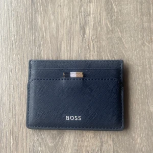 Svart kortfodral från BOSS - Snyggt och stilrent svart kortfodral från BOSS. Tillverkat i skinn med flera kortfack och en elegant detalj i metall. Perfekt för att hålla dina kort organiserade och lättillgängliga. Nypris 829kr.