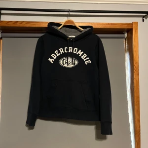  Abercrombie hoodie - Säljer en svart hoodie från Abercrombie med vit text och ett tryck på framsidan. Tröjan har en klassisk känguruficka och justerbar huva med snören. Perfekt för en avslappnad stil.