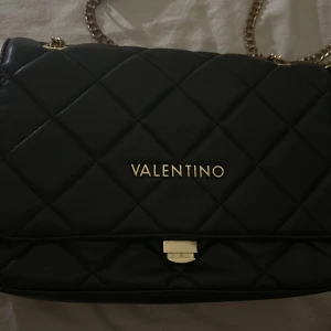 Svart quiltad axelväska från Valentino - Snygg svart axelväska från Valentino med quiltad design och guldfärgade kedjehandtag. Väskan har en elegant look med logotyp framtill och en praktisk dragkedja för säker förvaring. Ny väska 