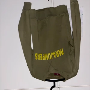 Olivgrön tröja från Parajumpers - Säljer en olivgrön tröja från Parajumpers med tryck i gult på framsidan. Tröjan har långa ärmar och en rund halsringning. Perfekt för en avslappnad stil.
