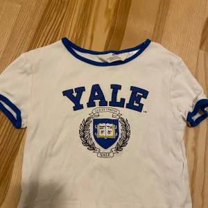 Vit t-shirt med Yale-tryck från H&M - Säljer en vit t-shirt från H&M med blå detaljer och ett stort Yale-tryck på framsidan. T-shirten är en kortare modell och säljes i fint skick! 