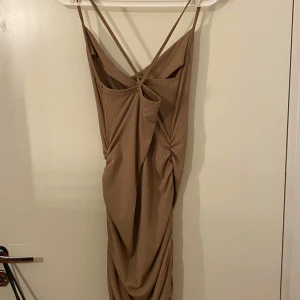 Beige klänning med drapering - Snygg beige klänning med draperad detalj på sidan. Klänningen har smala axelband som korsas i ryggen och en elegant omlott-look. Perfekt för en stilren och chic look.