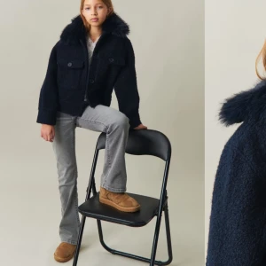 Mörkblå teddyjacka med pälskrage - Säljer en snygg mörkblå teddyjacka med en fluffig pälskrage. Jackan har en kort och boxig passform med knappar framtill och två fickor på bröstet. Perfekt för kyliga dagar när du vill vara både varm och stilren.