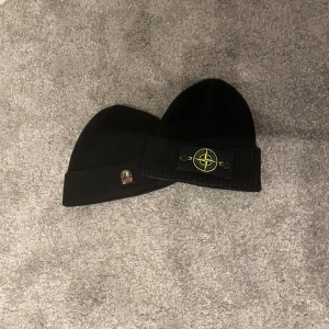 Svarta mössor från Parajumpers och Stone Island - Två svarta ribbade mössor, en från Parajumpers med broderad logotyp och en från Stone Island med den ikoniska kompasslogotypen. Perfekta för att hålla värmen med stil.