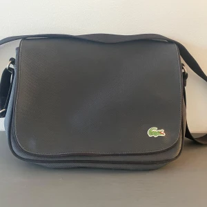 Svart axelväska från Lacoste - Snygg svart axelväska från Lacoste med klassisk krokodillogga. Väskan har ett randigt foder i grönt och beige samt flera praktiska fack med dragkedja. Perfekt för att hålla ordning på dina saker. Grå väska från Lacoste.   Använd, lite repig på locket i övrigt i fint begagnat skick.  Mått Bredd: ca 30 cm Höjd: ca 22 cm Djup: ca 6 cm  Kommer från ett djur och rökfritt hem.