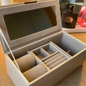 Beige smyckeskrin med spegel - Elegant beige smyckeskrin som aldrig använts med inbyggd spegel i locket. Skrinet har flera fack för förvaring av smycken, inklusive kuddar för armband och ringar. Perfekt för att organisera dina smycken på ett stilfullt sätt.  Bredd= 21 cm   Höjd= 8,5 cm