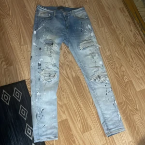 Blå jeans med slitningar från Amiri - Snygga blå jeans från Amiri med slitningar och färgstänk för en edgy look. De har en dragkedja och knapp framtill. Perfekta för dig som vill ha en unik stil.