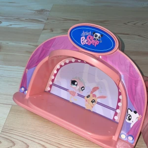 Littlest Pet Shop leksaksscen - Säljer en rosa leksaksscen från Littlest Pet Shop. Scenen har en söt design med två figurer i bakgrunden och en blå logotyp på toppen. Perfekt för att visa upp dina små figurer och skapa roliga berättelser.