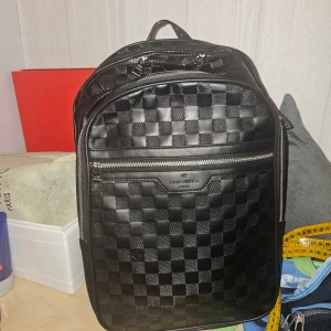 Svart rutig ryggsäck  - Snygg svart ryggsäck med klassiskt rutigt mönster. Väskan har flera dragkedjor och ett stilrent design. Perfekt för att bära med sig allt man behöver med stil.