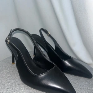 Svarta klackar - Eleganta svarta pumps med spetsig tå och smal klack. Skorna har en justerbar rem runt hälen för bättre passform. Aldrig använt dem, då de är precis för små för mig!!