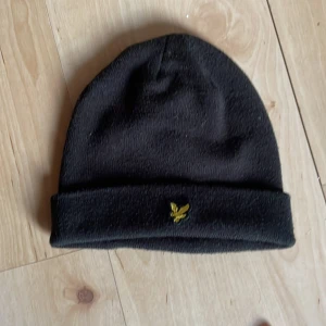 Svart mössa från Lyle & Scott - Snygg svart mössa från Lyle & Scott med ett litet gult broderat emblem framtill. Perfekt för kyliga dagar och enkel att matcha med olika outfits. Använd 1 gång pris kan diskuteras 