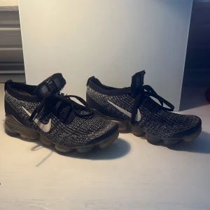 Nike Air Vapormax Flyknit sneakers - Snygga Nike Air Vapormax Flyknit sneakers i svart och grått. Skorna har en unik sula med synliga luftkuddar för extra komfort och stil. Ovandelen är i stickat material med snörning för en bra passform. Perfekta för både träning och vardag. Storlek 42 men passar allt mellan 39 och 43. Tveka inte att fråga vid minsta fundering. Pris kan diskuteras.
