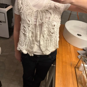 Vit stickad väst från Zara - Säljer en vit stickad väst från Zara med vackert virkat mönster och knytning framtill. Västen har en v-ringad design och är tillverkad i ett mjukt material. ALDRIG ANVÄND!!