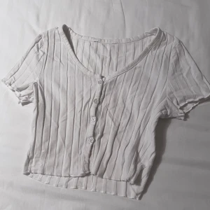 Vit ribbad kortärmad topp - Säljer en vit ribbad topp med korta ärmar och knappar framtill. Toppen har en v-ringad design och är croppad, vilket ger en trendig look. Kontakta mig om du har några frågor☺️