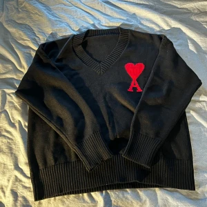 Svart tröja med rött hjärta från Ami Paris - Säljer en svart tröja från Ami Paris med ett stort rött hjärta och bokstaven A på bröstet. Tröjan har en v-ringad hals och ribbade muddar vid ärmslut och nederkant. Perfekt för en stilren look.