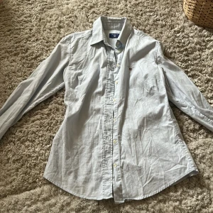 Ljusblå skjorta från GANT - Snygg ljusblå skjorta från GANT med klassisk krage och knappar framtill. Skjortan har ett broderat GANT-emblem på bröstet och långa ärmar. Perfekt för en stilren look.