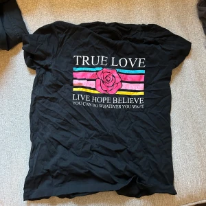 Svart t-shirt med tryck - Snygg svart t-shirt med färgglatt tryck och texten 'TRUE LOVE'. Perfekt för en avslappnad stil. T-shirten har korta ärmar och är gjord i ett mjukt material. Storlek xs-s